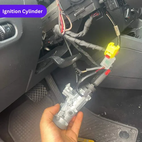 Photo of a hand holding a removed ignition cylinder from a car, showing the key slot and internal mechanism, with the dashboard and wiring visible in the background.