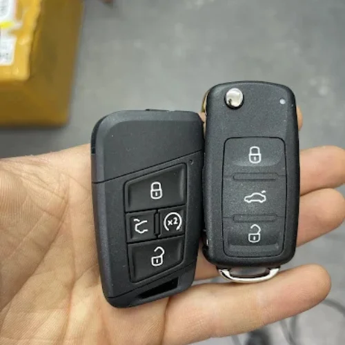 Hand holding two black car key fobs. Left one is a modern VW/Audi 5-button smart key (lock, unlock, trunk, x2 lock, panic), right one is a 4-button flip key with folded metal blade (lock, unlock, trunk, )