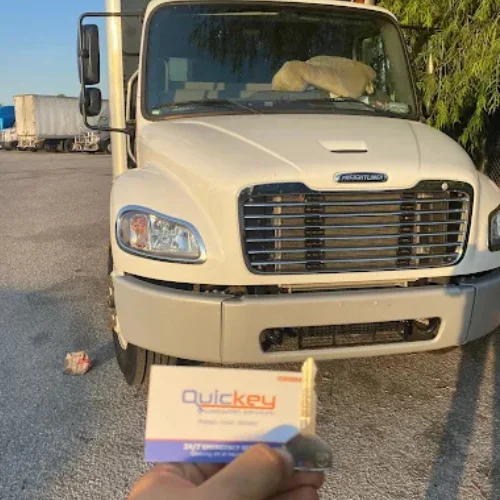 A person standing in front of a white Freightliner semi-truck, holding a newly replaced traditional metal ignition key with Quickey Locksmith Services business card after availing their on-site car key replacement service.