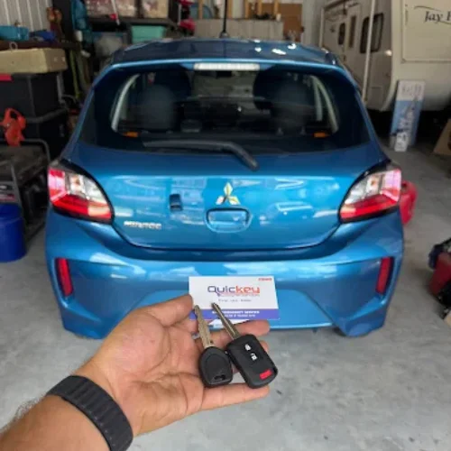 A person holding two car keys in hand – one traditional key and one transponder chip key. along with a Quickey Locksmith Services business card, in front of the rear view of a blue Mitsubishi Mirage hatchback parked in a garage.