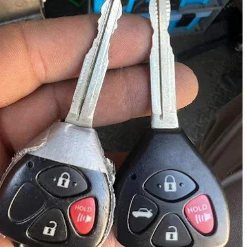 A hand holding two similar car key fobs side by side for comparison. Both have the same design black in color with four buttons (lock, unlock, trunk, and red panic button), but the left one has silver duct tape wrapped around its head.