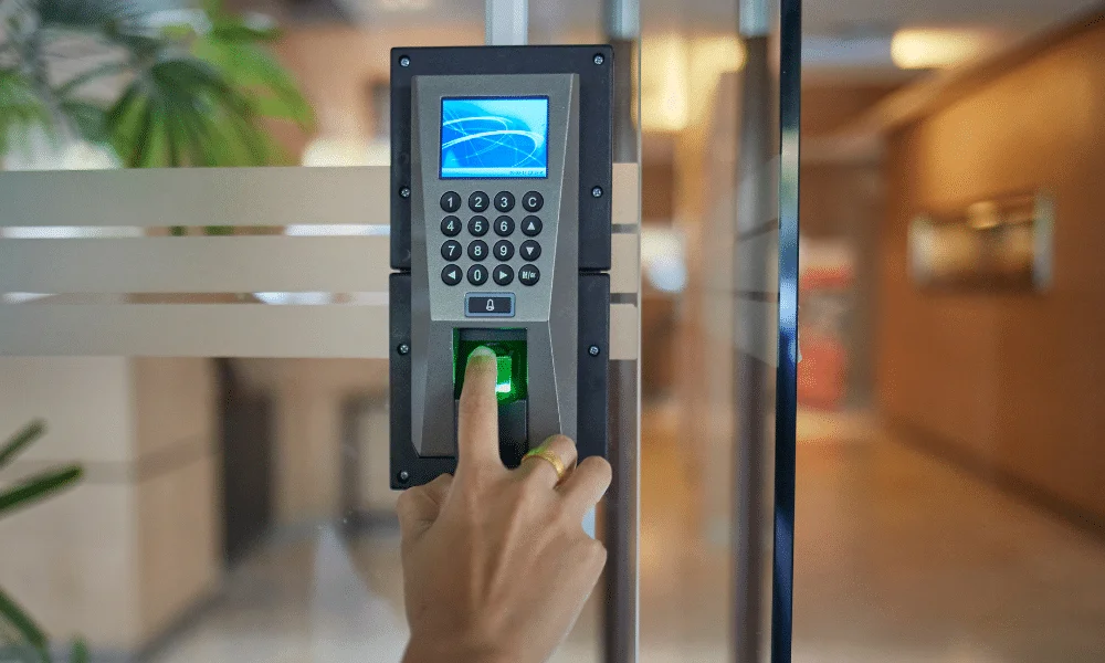 A person pressing their finger on a biometric fingerprint access control system installed on a glass commercial door. The system includes a digital screen and keypad, and is being used to securely unlock and control entry into a modern office or commercial building.