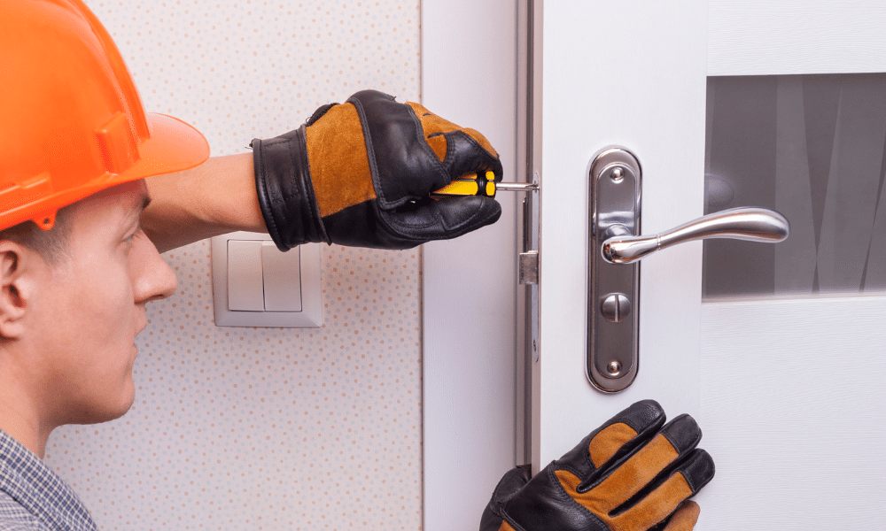 A professional locksmith wearing protective gloves and a helmet installing a door lock on a residential door using a screwdriver.