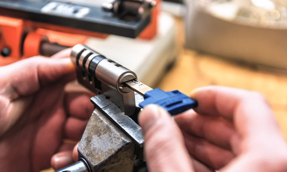 Locksmith hands adjusting a metal commercial door lock cylinder secured in a bench vise to test a blue-handled key.