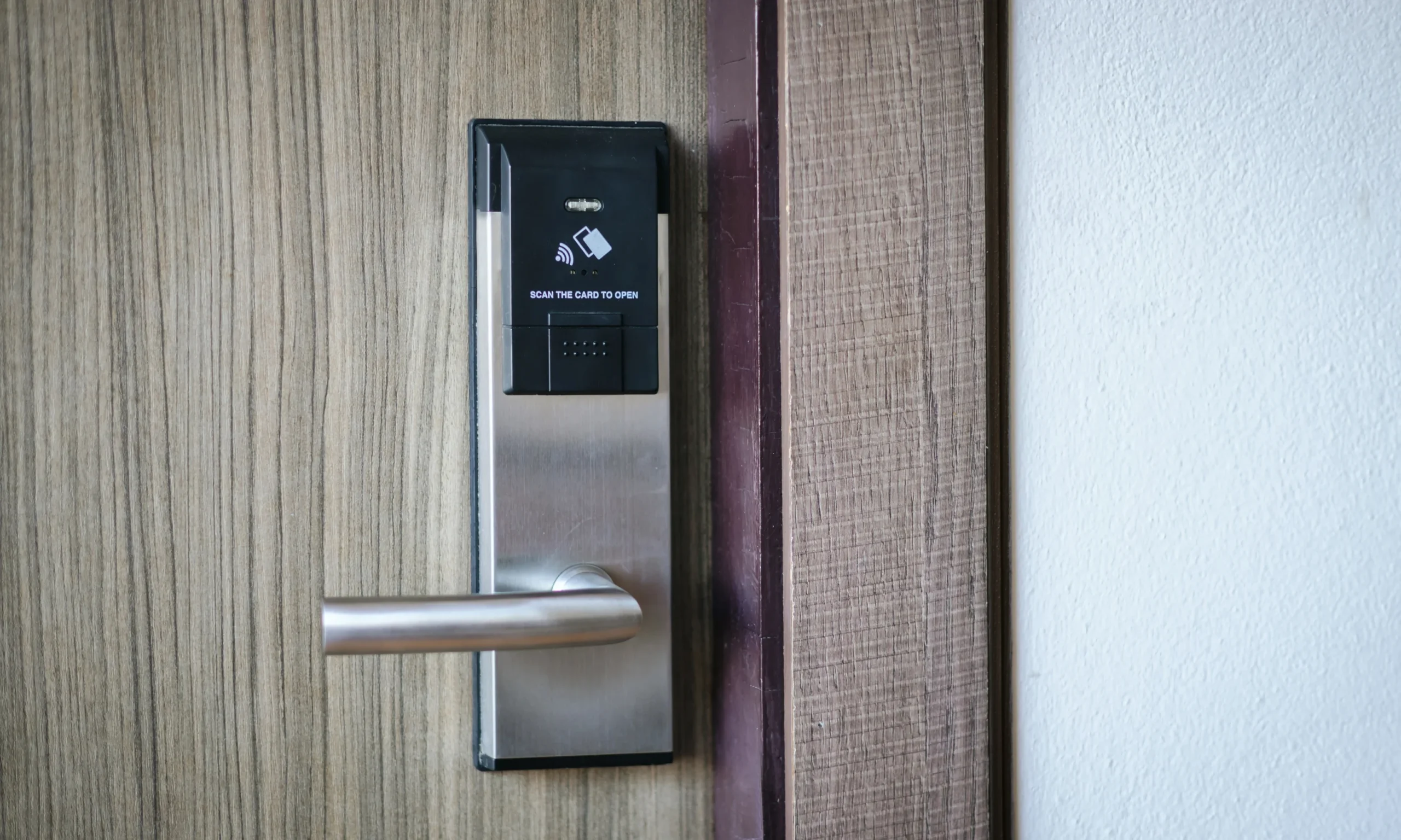 A close-up front view of a modern electronic door lock installed on a light wood-grain residential door, featuring a brushed stainless steel faceplate, matching horizontal lever handle, and a black RFID access panel mounted above the handle. The access panel displays a contactless card icon and the instruction "Scan the Card to Open," indicating support for secure key card entry. The surrounding wooden door frame and clean white interior wall create a neat, contemporary setting, while the minimalist design highlights the lock's advanced access technology. The image represents a fully configured electronic locking system with modern access features, reflecting the completed setup of a smart lock ready for secure everyday use through supported authentication methods.