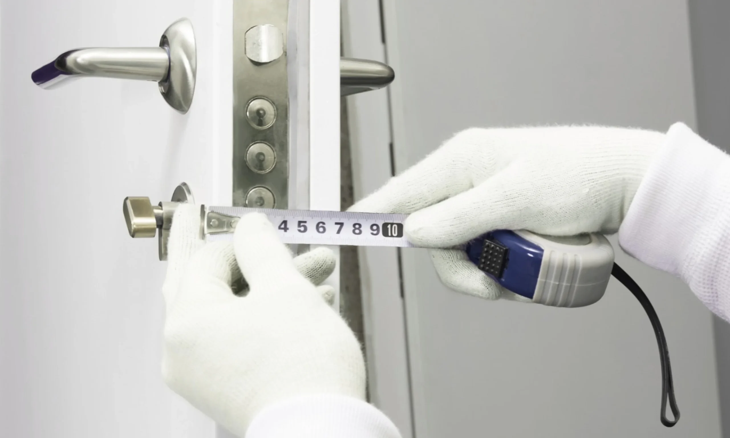 A close-up view of a technician wearing clean white protective gloves carefully measuring the dimensions of a residential door lock using a retractable measuring tape before installation. The white door is partially open, revealing its metal edge with multiple locking points, while a silver cylindrical lock component and brushed stainless steel lever handles are visible on both sides of the door. The measuring tape is precisely aligned across the lock hardware to verify spacing and fitment, emphasizing the importance of accurate door and lock measurements. The clean indoor setting, neutral background, and detailed focus on the hardware highlight a professional door compatibility check, ensuring the existing door and lock are properly assessed for a secure and compatible smart lock installation.
