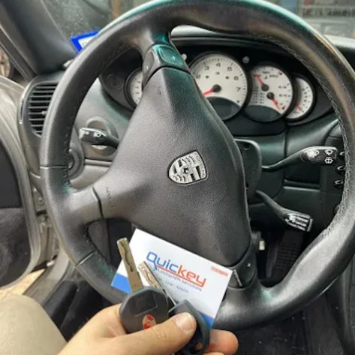 A close-up view from inside a vehicle showing a precision-cut laser-cut car key held in front of the steering wheel and instrument cluster. The replacement key features a high-security laser-cut blade with grooves machined into both sides for improved accuracy and compatibility with modern vehicle locking and ignition systems. A Quickey Locksmith Services key tag is positioned behind the key, while the dashboard gauges, steering wheel, and driver's area remain visible in the background, indicating the key is ready to be tested with the vehicle. The image represents a professional laser-cut and high-security key replacement service, providing a precisely cut replacement key designed to deliver reliable vehicle access, secure ignition operation, and compatibility with modern automotive security systems.