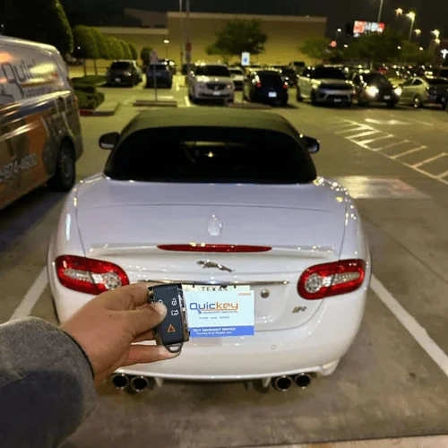A close-up view of a smart key fob held in front of a white luxury convertible parked in a well-lit parking lot at night. The black smart key features buttons for remote locking, unlocking, trunk release, and additional vehicle functions, allowing keyless entry and push-to-start operation on compatible vehicles. A Quickey Locksmith Services key tag is attached behind the smart key, while the rear of the vehicle, including its taillights, license plate area, and dual exhaust outlets, is visible in the background. Several parked vehicles, parking lot lighting, and a service van can also be seen, indicating the replacement was completed on-site. The image represents professional smart key replacement, providing a fully functional keyless entry and push-to-start key that restores secure, convenient access and reliable vehicle operation.