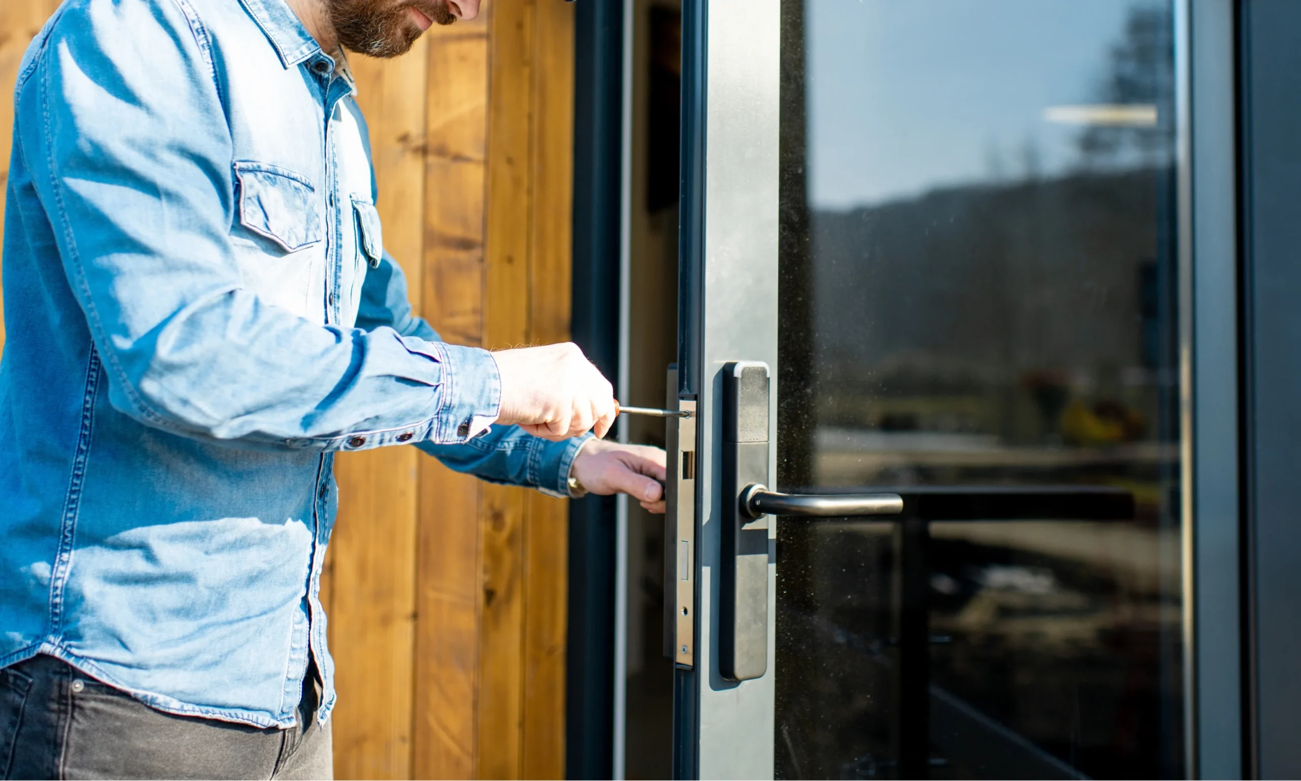 A close-up side view of a professional technician carefully installing a modern smart lock on a residential glass entry door using a handheld screwdriver. The technician, wearing a light blue denim work shirt, is precisely securing the lock mechanism while supporting the door hardware with the other hand to ensure proper alignment and a secure fit. The contemporary door features a black metal frame, a sleek matte black smart lock with a matching horizontal lever handle, and a large glass panel reflecting the outdoor surroundings. Warm natural wood paneling beside the entrance contrasts with the dark door frame, while bright daylight illuminates the installation process, highlighting the craftsmanship and attention to detail. The image represents professional smart lock installation on a compatible residential door, emphasizing accurate fitting, secure hardware installation, and reliable home security for modern keyless entry systems.
