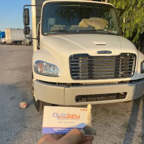 A close-up view of a traditional metal car key held in front of a white commercial truck parked outdoors. The classic single-sided metal key is attached to a Quickey Locksmith Services key tag and is positioned in the foreground, while the truck remains slightly blurred in the background to emphasize the key. The vehicle features a large front grille, wide windshield, and heavy-duty design, representing an older model that commonly uses traditional mechanical keys instead of electronic key fobs or smart keys. The image illustrates a professional traditional car key replacement service, showing a newly cut replacement key ready for use with a compatible vehicle, helping restore reliable access and ignition for older cars and trucks.