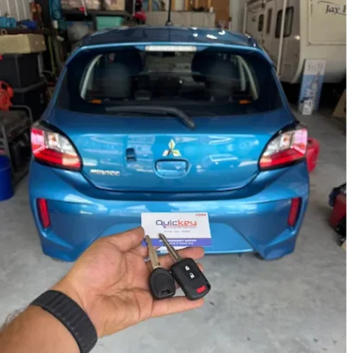 A close-up view of a person's hand holding a newly cut transponder chip key and its matching remote key fob in front of a blue Mitsubishi hatchback parked inside a garage. The transponder key features a built-in security chip within the key head, designed to communicate with the vehicle's immobilizer system for authorized starting. A Quickey Locksmith Services key tag is positioned behind the keys, while the rear of the vehicle remains slightly blurred in the background to keep the focus on the replacement keys. The garage setting includes automotive equipment and storage items, indicating the work is being completed by a professional locksmith. The image represents professional transponder chip key replacement, providing a properly cut and programmed key that restores secure vehicle access and reliable engine starting for compatible vehicles.