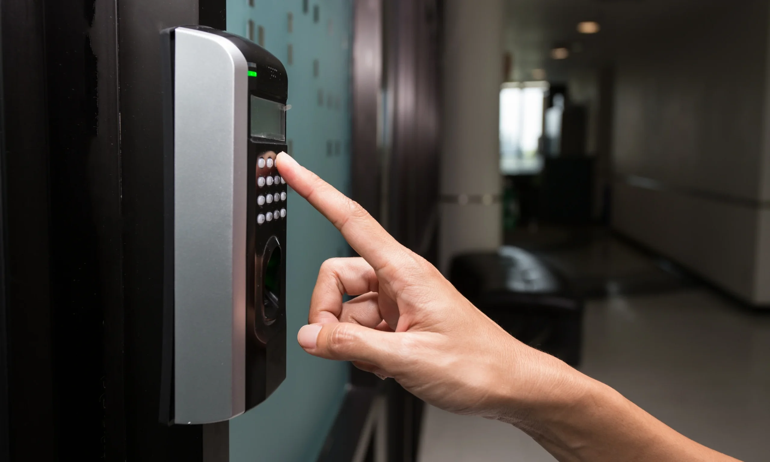 A close-up side view of a person using their index finger to enter a code on the numeric keypad of a modern electronic access control device mounted beside a glass entry door. The slim black and silver unit features a digital display, illuminated status indicator, numeric keypad, and an integrated fingerprint scanner positioned below the buttons. The surrounding entrance has a dark metal door frame with frosted glass panels, while the softly blurred indoor hallway in the background creates a clean, professional environment. The image highlights the process of configuring and using secure access credentials, including PIN code entry and biometric authentication, demonstrating the personalized access features available on supported electronic locking systems.