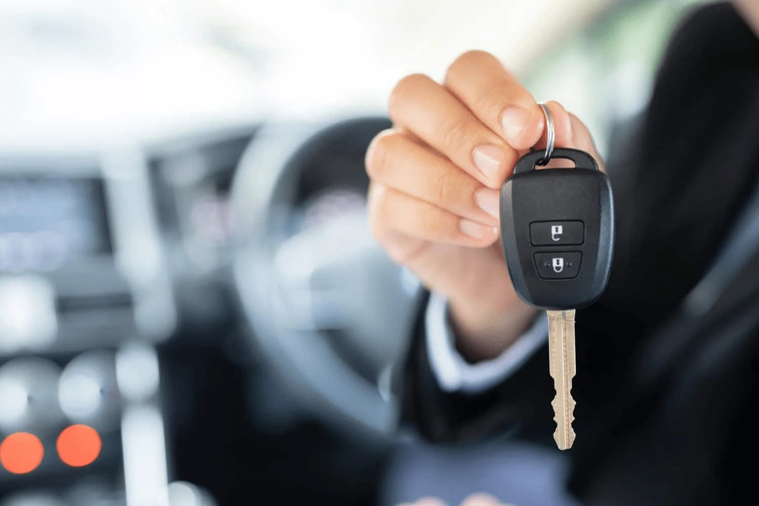 A close-up view inside a vehicle showing a person holding a black automotive key fob with an attached metal car key blade. The key fob is held prominently in the foreground by a person wearing a dark suit or jacket, with the fingers wrapped around a small metal key ring at the top of the fob. The black plastic key fob has two clearly visible rectangular buttons on its front, each marked with a small white lock symbol, representing vehicle locking and unlocking functions. A traditional metal key blade extends downward from the bottom of the key fob, with its cut edges and grooves visible along the length of the blade. The key is positioned in front of the vehicle's interior, while the steering wheel, dashboard, center console, and other interior components appear softly blurred in the background. The shallow depth of field keeps the automotive key and remote fob as the main focus of the image. The scene visually represents automotive locksmith services involving car key replacement, key fob replacement, car key duplication, and programming of vehicle keys and remotes, while also relating to services such as transponder key programming and assistance with vehicle ignition-related key issues. The image supports Quickey Locksmith Services automotive locksmith services for customers in Alief, Houston, TX.