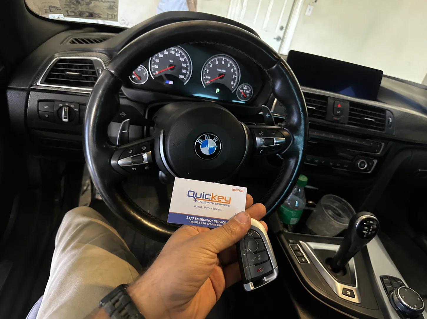 A customer holding a newly prepared BMW smart key fob alongside a Quickey Locksmith Services business card inside a BMW vehicle. The black BMW smart key fob features multiple integrated buttons, a silver trim edge, and a compact rectangular design for the vehicle’s electronic locking and security functions. The BMW steering wheel with its roundel emblem, illuminated instrument cluster, dashboard, center console, gear selector, and infotainment controls are visible in the background. The Quickey Locksmith Services business card is positioned beside the BMW smart key fob, showing the company logo and locksmith service information. The image represents a completed BMW car key replacement and programming service provided by Quickey Locksmith Services to a customer in Clear Lake, Houston, TX.