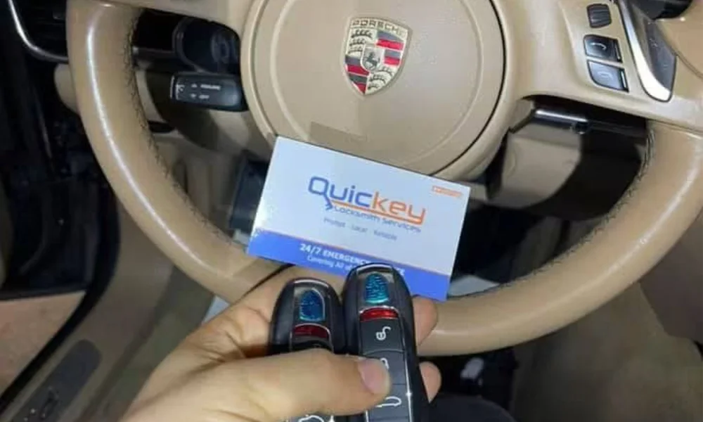 Customer review photo showing an old Porsche key fob that was no longer working alongside a newly duplicated Porsche key fob made by Quickey Locksmith Services, both held in one hand in front of a beige Porsche steering wheel. The two black key fobs feature Porsche-style buttons and colored button markings, with the older non-working key positioned beside the newly made replacement key. Behind the keys, the beige leather steering wheel prominently displays the Porsche crest in the center, with steering-wheel controls visible on the right side and dashboard controls visible behind the wheel. A Quickey Locksmith Services business card is held behind the key fobs, displaying the company name in blue and orange lettering, contact information, and a blue strip identifying 24/7 emergency service. The customer provided the photo as a review after having a new duplicate Porsche car key made by Quickey Locksmith Services to replace the old key that was no longer functioning. The image represents professional automotive locksmith services in Conroe, TX.