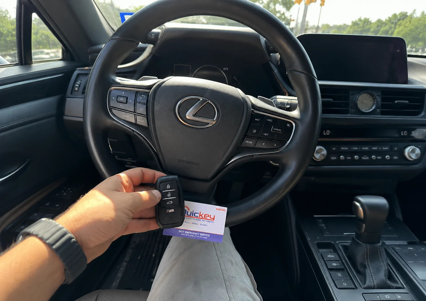 A customer holding a newly prepared Lexus smart key fob alongside a Quickey Locksmith Services business card inside a Lexus vehicle. The black Lexus smart key fob has multiple integrated buttons for locking, unlocking, and opening the vehicle trunk, with the Lexus logo visible on the steering wheel. The Lexus steering wheel and emblem, instrument cluster, dashboard, infotainment display, climate controls, center console, and automatic gear selector are visible in the vehicle interior. The Quickey Locksmith Services business card is held behind the key fob, showing the company branding and automotive locksmith service information. The image represents a completed Lexus car key replacement and programming service provided by Quickey Locksmith Services to a customer in Downtown Houston, TX.