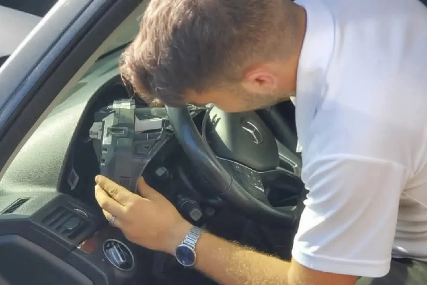 Automotive locksmith in Kingwood, Houston, TX, reinstalling the removed steering wheel and steering-column trim inside a black Mercedes vehicle after completing the locksmith work behind the steering area. The locksmith is leaning into the driver’s side of the car and carefully holding the black plastic steering-column panel in place with both hands, aligning it around the steering wheel and instrument cluster. He is wearing a white short-sleeve shirt and a silver wristwatch. The Mercedes steering wheel with its emblem, black dashboard, instrument cluster, air vents, center console, and driver-side controls are visible throughout the vehicle interior. The removed panel exposes the area behind the steering wheel where the repair was completed, while the locksmith is putting the surrounding section back into its original position. Natural daylight illuminates the vehicle interior. The image illustrates professional automotive locksmith services provided by Quickey Locksmith Services in Kingwood, Houston, TX.