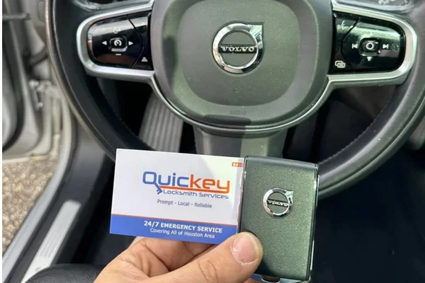 A customer-submitted photo showing a repaired Volvo key fob and Quickey Locksmith Services business card inside a Volvo vehicle. The black steering wheel is prominently visible in the background with the Volvo logo in the center, along with control buttons on both sides and part of the vehicle’s dashboard and driver-side interior. In the foreground, a hand is holding a rectangular black Volvo key fob with the Volvo logo visible on its front surface. Beside and partially behind the key fob is a white Quickey Locksmith Services business card featuring the blue and orange Quickey logo, the words “Locksmith Services,” the tagline “Prompt • Local • Reliable,” and a blue section at the bottom stating “24/7 EMERGENCY SERVICE” and “Covering All of Houston Area.” The vehicle interior is primarily black and gray, with the driver-side door area visible on the left and the steering wheel centered behind the key and business card. The photo was provided by a customer as a review after Quickey Locksmith Services repaired the customer’s Volvo key fob and returned the working key to the owner. The image represents automotive locksmith services in League City, Texas.