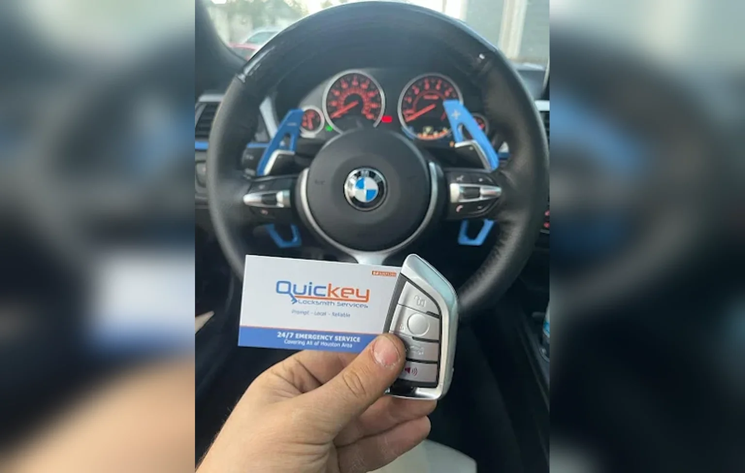 A customer holds a newly made silver-and-black BMW key fob and a Quickey Locksmith Services business card inside a BMW, with the BMW logo clearly visible in the center of the black steering wheel and the vehicle’s illuminated instrument cluster visible behind it. The key fob is held in the lower-right foreground and has a silver outer frame with several black and silver buttons, including the lock, unlock, and other vehicle-control buttons. The business card is held beside the key fob and displays the “Quickey Locksmith Services” logo in blue and orange, the tagline “Prompt • Local • Reliable,” and a blue bottom section reading “24/7 EMERGENCY SERVICE” and “Covering All of Houston Area.” The black steering wheel surrounds the center of the image, with blue paddle-shift accents visible behind the wheel on both sides. Red-lit gauges and indicators can be seen on the dashboard through the steering wheel, while the customer’s hand holds the new key fob and business card in the foreground, illustrating a completed automotive locksmith service for a BMW customer in Pasadena, Texas.