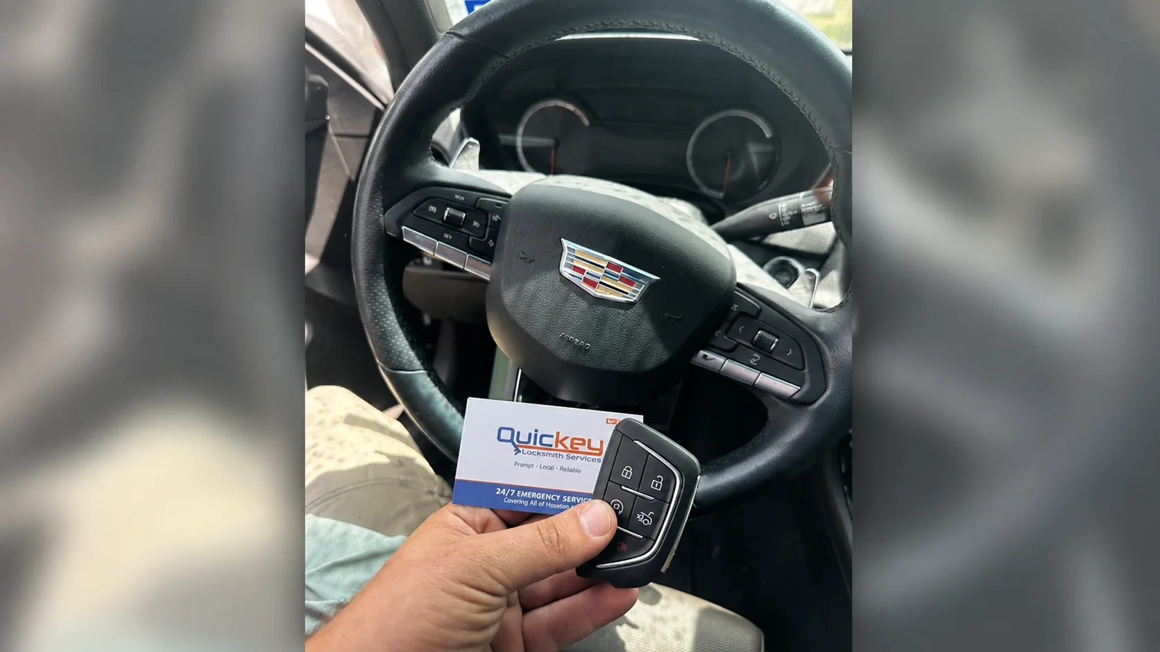 A customer photo taken inside a Cadillac vehicle in Pearland, TX, showing a newly made Cadillac key fob alongside a Quickey Locksmith Services business card. The center of the image is dominated by the vehicle’s black leather-wrapped steering wheel, with the Cadillac crest emblem clearly visible on the center airbag cover. Behind the steering wheel, the dark instrument cluster contains two circular gauge displays, while the steering-wheel spokes have integrated black control buttons and silver trim. A turn-signal and control stalk extends from behind the right side of the steering wheel. In the lower center of the image, a person’s hand is holding a black Cadillac key fob in front of the steering wheel. The key fob has a rounded rectangular shape with a silver metallic edge and several clearly visible buttons, including lock, unlock, and other vehicle-control buttons. A metal key blade extends from the lower side of the fob. Behind and partially beneath the key fob, the person is also holding a Quickey Locksmith Services business card. The card has a white background with the “Quickey” name in blue and orange lettering, the words “Locksmith Services” underneath, and additional small company text and service information printed below. The lower portion of the card has a blue section with white lettering. The person’s forearm and hand are visible in the foreground, with light-colored clothing and beige pants visible around the driver’s seat area. The Cadillac interior is predominantly black, including the steering wheel, dashboard, controls, and surrounding trim. The image is vertically framed with blurred portions of the same vehicle scene along both outer sides. The photo represents a customer displaying the newly made Cadillac key fob after receiving automotive locksmith service from Quickey Locksmith Services in Pearland, TX.