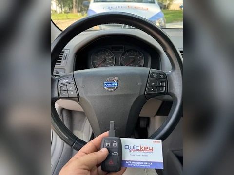 A customer photo taken inside a Volvo vehicle showing a repaired Volvo slot remote key and a Quickey Locksmith Services business card held in front of the steering wheel. The black steering wheel has the Volvo logo clearly visible in the center, with the instrument cluster and dashboard controls visible behind it. In the foreground, a hand is holding a black Volvo remote key with visible lock, unlock, and red alarm buttons. The Quickey Locksmith Services business card is held beside the key and shows the company logo and service information. Through the windshield, part of a Quickey Locksmith Services van is visible outside. The image represents mobile automotive locksmith service provided by Quickey Locksmith Services in Spring, TX.