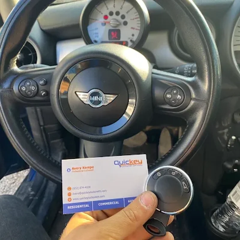 A customer-submitted photo showing a Mini Cooper interior after receiving automotive locksmith service in Sugar Land, Texas. The image is taken from the driver’s seat area and prominently shows the black leather steering wheel with the MINI winged logo in the center, along with the vehicle’s circular instrument cluster, dashboard controls, steering-wheel buttons, and ignition area. In the lower center of the image, a hand is holding a black and silver Mini Cooper key fob. The key fob is positioned directly in front of the steering wheel and ignition area. A Quickey Locksmith Services business card is also held in front of the steering wheel, partially underneath the key fob. The business card features the Quickey Locksmith Services logo near the upper right and printed contact information, with colored service labels along the bottom including “RESIDENTIAL,” “COMMERCIAL,” and “AUTOMOTIVE.” The vehicle interior is predominantly black and dark gray, with the steering wheel, dashboard, instrument panel, control buttons, and ignition area clearly visible. The photo documents a Mini Cooper key and Quickey Locksmith Services business card following automotive locksmith service and represents automotive locksmith services in Sugar Land, Texas, particularly Mini Cooper key and key fob service.