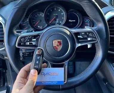 A customer-submitted photo showing a Porsche car interior with a replacement Porsche key fob and Quickey Locksmith Services business card displayed in front of the steering wheel. The black leather steering wheel has the Porsche crest in the center and multiple control buttons on both sides. Behind the steering wheel, the dashboard features several round instrument gauges, including the central tachometer and additional gauges on either side. A hand in the lower-left portion of the image is holding a black Porsche key fob upright, with several visible buttons on its front. A Quickey Locksmith Services business card is positioned directly behind the key fob at the bottom center of the image, partially visible beneath the hand and key. The visible portion of the card includes the Quickey Locksmith Services branding and blue-colored design elements. The image focuses on the Porsche steering wheel, vehicle instrument cluster, replacement key fob, and Quickey Locksmith Services business card, representing automotive locksmith services in The Woodlands, Texas and a completed Porsche key fob service.
