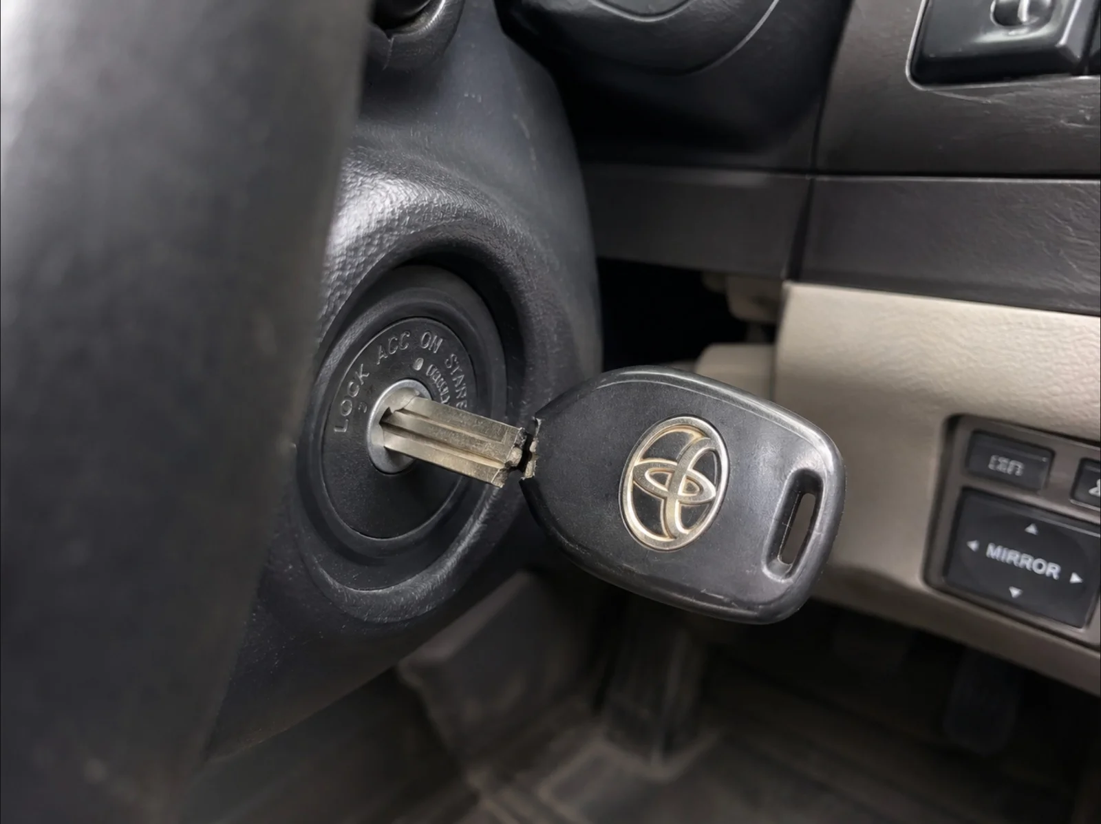 A close-up view of a broken Toyota mechanical key inserted into the vehicle's ignition cylinder inside the driver's area. The metal key blade remains inserted in the ignition, while the black plastic key head with the silver Toyota logo is visibly cracked and separated at the base where it connects to the blade. The ignition switch displays the LOCK, ACC, ON, and START positions around the key cylinder. Part of the steering column, dashboard controls, and the driver's footwell are visible in the background, providing context for the vehicle interior. The image represents a broken Toyota key that has failed while inserted in the ignition, requiring professional key extraction and replacement to restore normal vehicle operation.