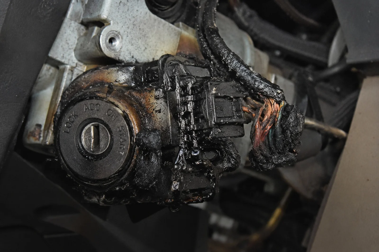 A close-up view of a severely burnt vehicle ignition switch assembly mounted inside the exposed steering column. The circular ignition cylinder is visible on the left with the positions "LOCK," "ACC," "ON," and "START" clearly marked around the key slot. Attached to the back of the ignition switch is a heavily melted black electrical connector covered with thick burned residue. The surrounding wiring harness has extensive fire damage, with the protective insulation melted away and several copper wires exposed, twisted together, and darkened from extreme heat. Burn marks, soot, and melted plastic are visible across the ignition switch housing, electrical connector, and surrounding components, showing significant overheating and electrical failure. The interior steering column structure and metal mounting bracket are partially visible in the background, while the focus remains on the damaged ignition switch and burnt wiring. The image illustrates a dangerously burnt ignition switch that has suffered severe electrical damage and requires professional ignition switch replacement to restore safe and reliable vehicle operation.