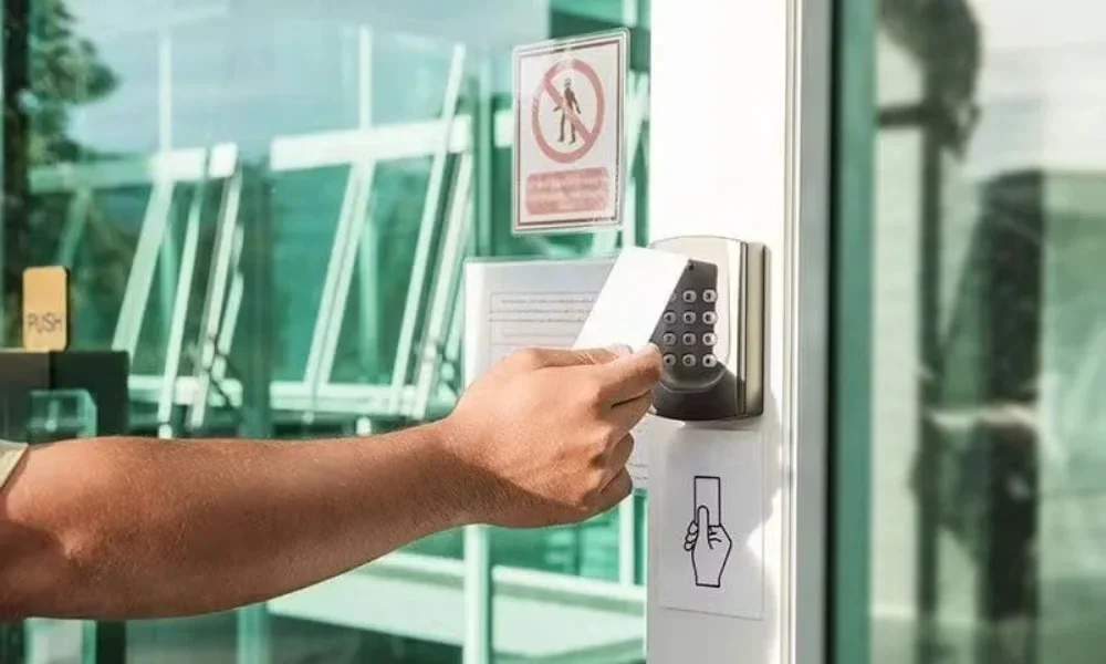 Commercial locksmith services in Conroe, TX represented by an electronic access-control system installed beside a glass commercial entrance. A person’s arm and hand extend from the left side of the image toward a wall-mounted access-control reader, holding a white access card directly in front of the reader as if presenting the card to unlock or gain authorized entry through the door. The access-control unit is mounted vertically on a silver metal plate and features a black numeric keypad with multiple round buttons arranged in rows, along with a small reader area near the top. The reader is attached to the white vertical frame of a glass entrance door. Below the access-control unit, a printed instructional sign shows a hand pressing or using the door-access area. Behind the entrance, large glass panels and windows reveal a bright commercial interior or building entry area, with reflections and structural window frames visible through the glass. A red-and-white warning or restriction sign is posted on the glass above and slightly behind the access-control reader. The scene is brightly illuminated with natural daylight, and the glass surfaces create greenish reflections throughout the background. The image illustrates commercial locksmith services for businesses in Conroe, TX.