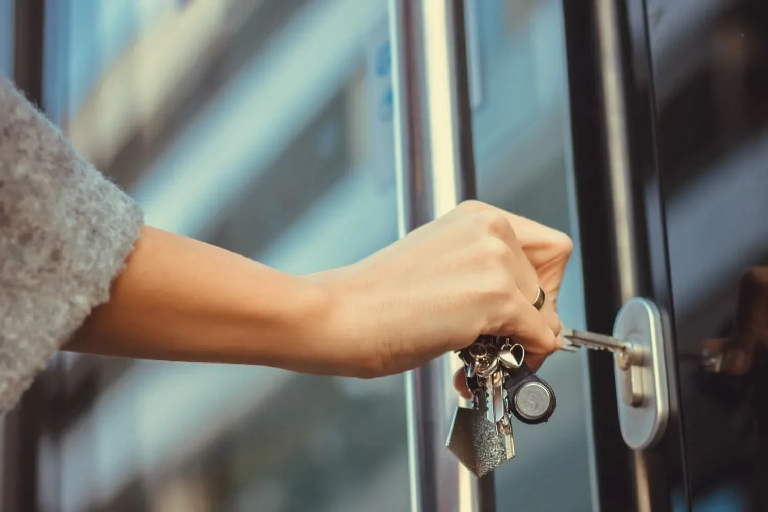 A close-up of a person using a set of keys to unlock a glass commercial door, with the key inserted into the exterior door lock while holding several keys on a keyring. The image shows the door handle, lock cylinder, glass panels, and the person’s hand turning the key, illustrating commercial locksmith services in Kingwood, Houston, TX.