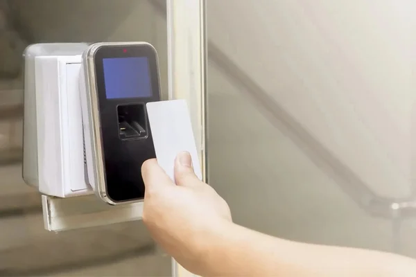 A close-up view of a commercial access control system installed beside a glass door. The wall-mounted electronic lock has a black front panel surrounded by a silver-colored frame and features a small blue display screen at the top, a biometric fingerprint scanner below the screen, and a small indicator light near the upper edge. A person’s hand is holding a white access card directly in front of the fingerprint scanner, demonstrating the use of an electronic access credential with the locking system. The access control device is mounted next to a white rectangular wall switch or electrical panel. The glass door and its vertical frame are visible beside the device, with a light-colored interior area seen through the glass. The image focuses on the electronic locking device, fingerprint scanner, display screen, access card, and door entry area. The image represents commercial locksmith services in League City, Texas, particularly commercial access control and electronic security system installation or service.
