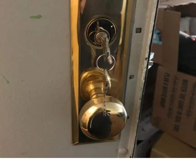 Close-up of a commercial storage room door with a white surface and brass-colored lock hardware, including an upper deadbolt and round doorknob. A metal key is visibly stuck inside the upper lock, with a key ring and additional keys hanging downward in front of the lock. The door is partially open, revealing cardboard storage boxes and the dim interior of the storeroom behind it.
