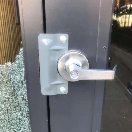 Close-up view of a commercial entrance door in Sugar Land, Texas, showing a silver metal lever door handle and lock mounted on a dark gray metal-framed door. The handle has a round silver lock housing with a key cylinder at its center and a horizontal rectangular lever extending toward the right. The lock and handle are attached to a large rectangular silver metal mounting plate secured to the door with visible screws. On the left side of the door, a large section of glass is broken, with numerous small shards and jagged pieces of shattered glass still visible along the edge of the opening. The broken glass contrasts with the intact dark reflective glass panel on the right side of the door, which reflects part of the surrounding outdoor area. The dark gray metal door frame and vertical seams are visible around the lock hardware, while a narrow strip of brown vertical paneling or wall material appears at the far left edge of the image. The close-up focuses on the commercial door hardware, locking mechanism, metal frame, and damaged glass, representing commercial locksmith service in Sugar Land, Texas.