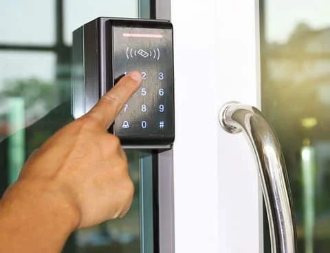 A close-up photograph of a commercial glass entrance door with a black electronic access control lock mounted beside the door. A person’s hand is pressing a number on the illuminated black touchscreen keypad, which displays a numeric keypad with digits, an access symbol near the top, and small lock icons near the bottom. The electronic lock has a rectangular black housing with a slightly reflective surface and is mounted on the dark metal frame of the glass door. To the right, the white door panel has a curved silver metal pull handle. Reflections of the surrounding building and outdoor area are visible through the glass, with soft natural daylight illuminating the scene. The image represents commercial locksmith service in Spring, TX.