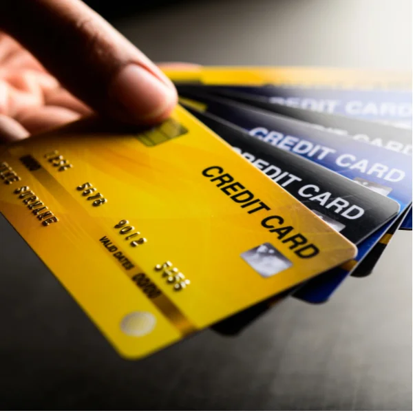 A close-up photograph of several credit cards stacked and partially overlapping on a dark surface. The front card is bright yellow with the words “CREDIT CARD” printed in bold black letters, along with a visible chip, card number, expiration date, and other standard card details. Several black, blue, and gray credit cards are layered behind it, with “CREDIT CARD” text visible on some of the cards. A hand is holding the upper edge of the yellow card near the top left of the image. The image visually represents credit card payment as a payment method.
