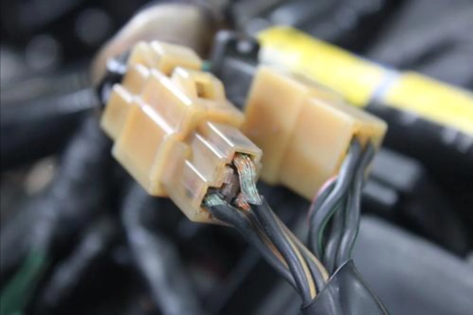 A close-up view of a damaged electrical connector attached to a vehicle's ignition switch wiring harness, showing exposed copper wires protruding from the plastic connector due to worn or broken insulation. Multiple black electrical wires extend from the connector, with the blurred engine or dashboard components visible in the background. The image illustrates a faulty ignition switch electrical connection that can cause intermittent starting problems, loss of power, or unreliable ignition operation and may be repaired without replacing the entire ignition switch.