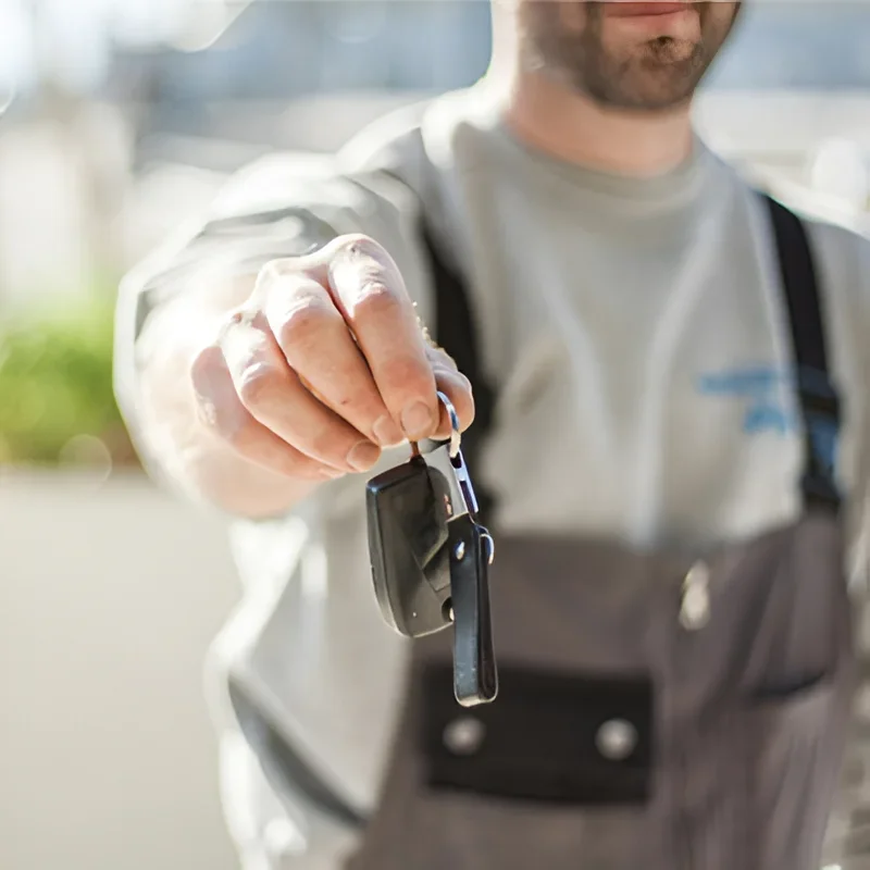 A close-up view of a professional locksmith handing a newly prepared car key toward a customer. The locksmith is wearing a light-colored professional work uniform with a dark utility pouch around his waist, and his face is partially cropped and softly out of focus near the top of the image. His extended hand is the main focus of the photograph, holding a black automotive key fob and attached metal key blade by a small key ring. The black key fob has a compact rectangular design with visible buttons and a separate metal key blade extending downward from it. The newly prepared vehicle key is presented prominently in the foreground, positioned between the locksmith and the person receiving the service. The locksmith is standing outdoors in bright natural daylight, with the background softly blurred to keep attention on the key and the handover. Greenery and a light-colored building or residential surroundings are faintly visible behind him, creating the appearance of an on-site mobile locksmith service visit. The shallow depth of field emphasizes the locksmith’s hand, the automotive key fob, and the metal key while leaving the locksmith and surrounding environment less defined. The scene represents a professional automotive locksmith completing a car key replacement for a customer after the original vehicle key was lost. The newly prepared key is being handed over after the locksmith has completed the replacement service, helping the customer regain access to and use of their vehicle. The image supports Quickey Locksmith Services Emergency Locksmith Services in Katy, TX, particularly situations involving lost vehicle keys and urgent automotive lock and key assistance.