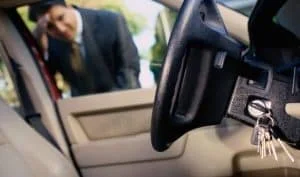 A close-up interior view of a vehicle showing the driver’s side area with the steering wheel, dashboard, ignition, and a set of metal car keys inserted into the ignition switch. The keys hang downward from the ignition near the steering column, while the black steering wheel and part of the vehicle’s dashboard and instrument panel are clearly visible in the foreground. The driver-side door is open, revealing the light-colored interior door panel and allowing a view toward the outside of the vehicle. A person wearing dark black pant suit is standing just outside the locked driver-side door and leaning toward the vehicle, appearing to assist with or inspect the vehicle from outside. The person is slightly out of focus while the steering wheel, ignition, and keys remain the primary visual focus. Natural daylight enters through the open door and windows, illuminating the vehicle’s interior. The image visually represents professional emergency automotive locksmith assistance, such as helping a customer regain access to a vehicle during a car lockout or other urgent vehicle access situation, and supports the Emergency Locksmith Services offered by Quickey Locksmith Services in Alief, Houston, TX.