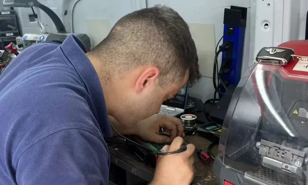 A locksmith working at a compact mobile workbench inside a service vehicle, carefully working on a green electronic circuit board with a small handheld tool connected by a black cable. The locksmith is leaning closely over the workbench and concentrating on the exposed circuit board, with one hand holding the tool and the other hand positioned near the board. He is wearing a dark blue short-sleeve polo shirt and has short brown hair. The work area contains small locksmith and electronic repair supplies, including a spool of solder, wires, hand tools, and other small components scattered around the bench. A large red-and-gray key-cutting or locksmith machine with a transparent protective cover occupies the right side of the image, while additional equipment, cables, and storage areas are visible around the vehicle interior. The surrounding walls and equipment are light gray and white, with a blue vertical power or electrical unit mounted toward the back. The image shows a locksmith performing detailed hands-on repair work with electronic components and specialized equipment as part of emergency locksmith service in Conroe, TX.