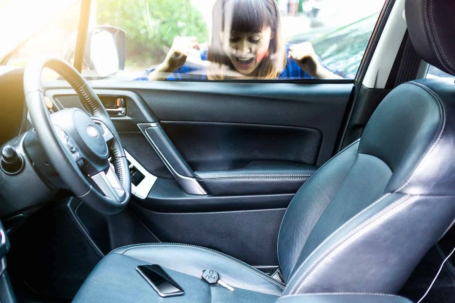 A view from inside a parked car showing a young girl standing outside the closed driver-side. The girl has straight brown hair with bangs and is wearing a blue plaid shirt. She is looking into the vehicle through the window with her mouth open and both arms raised near her shoulders, appearing concerned. The car interior is predominantly black and dark gray, with a black steering wheel on the left, a dark dashboard, the driver-side door panel, a large black front passenger seat on the right, and part of the center console visible. A black smartphone is lying on the front passenger seat next to a small black car key fob with several visible buttons. The driver-side window is closed between the girl and the vehicle interior, clearly separating her from the car. Bright daylight enters through the windows, and trees, greenery, and a vehicle are visible outside in the softly blurred background. The image focuses on the person outside the closed vehicle, the car interior, and the visible key fob left inside the car. The scene represents emergency locksmith services in Kingwood, Houston, Texas, particularly assistance with situations where a customer needs urgent access to a vehicle.