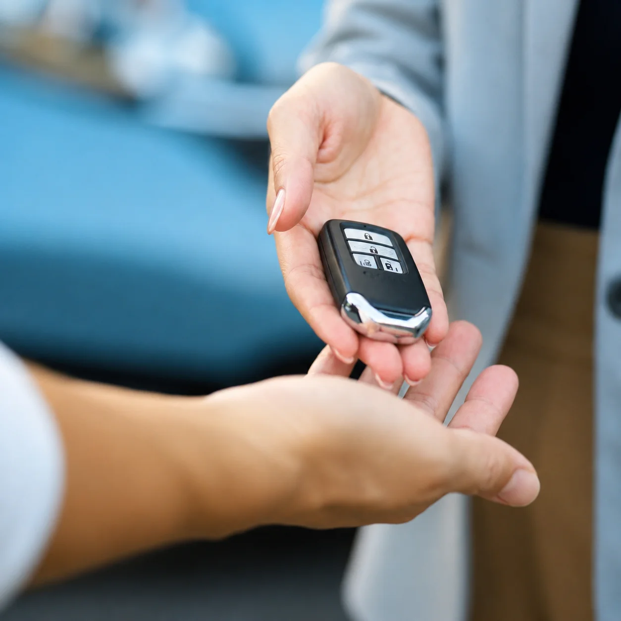 A close-up image showing a locksmith handing a newly made black car key fob to a customer in Sugar Land, Texas, after an emergency automotive locksmith service. The locksmith’s hands are positioned above and behind the key fob, carefully presenting it to the customer, while the customer’s hands are open underneath and around the fob as they receive it. The new key fob is black with silver-colored trim along its lower edge and has several rectangular buttons on its upper surface, including visible lock, unlock, and other vehicle-control symbols. The key fob is the main focus of the image and is held between the locksmith and customer during the handover. Only the hands and forearms of the two people are visible; their faces are outside the frame. The background is softly blurred and includes blue vehicle interior area, along with light gray and white surfaces, keeping attention on the key fob and the handover. The image represents an emergency locksmith service in Sugar Land, Texas, where a locksmith has provided a replacement car key fob after the customer’s original key fob was damaged or broken and was no longer usable.