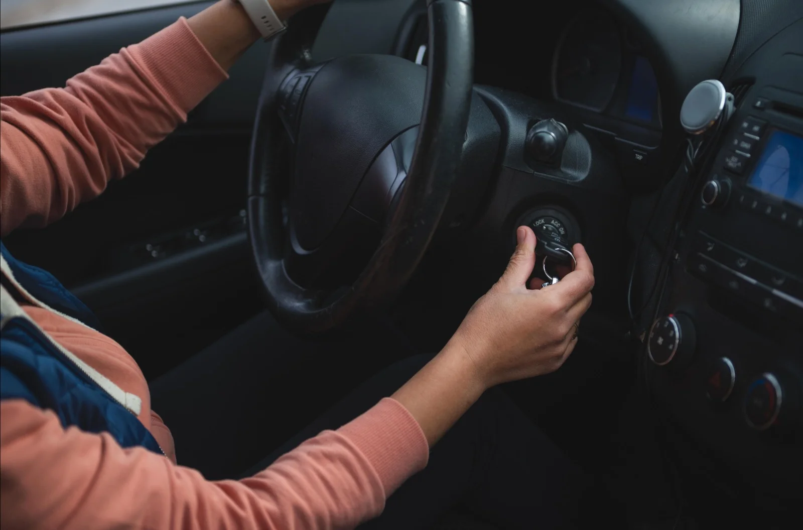 A close-up interior view of a person seated in the driver's seat of a vehicle while attempting to turn the key in the ignition switch. One hand grips the steering wheel, while the other hand holds a mechanical car key inserted into the ignition cylinder, positioned between the LOCK, ACC, ON, and START settings. The black steering wheel, instrument cluster, dashboard controls, center console, climate control knobs, and infotainment system are visible, providing a clear view of the driver's area. The person is wearing a long-sleeve peach-colored shirt and a smartwatch, with their posture focused on operating the ignition. Soft natural daylight illuminates the vehicle interior, emphasizing the ignition switch and key as the main focal point. The image represents a vehicle experiencing intermittent starting issues, where the ignition switch may require professional diagnosis and repair to restore reliable engine starting.
