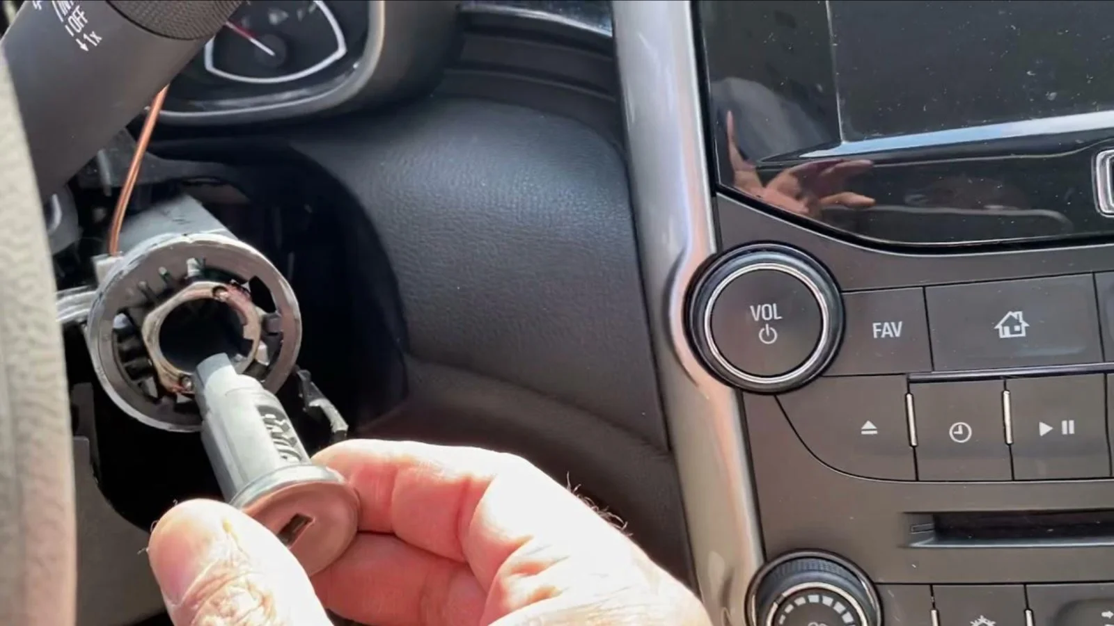A close-up view inside a vehicle showing a partially disassembled steering column with the ignition switch assembly exposed. A locksmith's hand is holding the metal ignition lock cylinder while carefully aligning it with the opening in the steering column, where the surrounding trim has been removed to access the internal ignition components. The exposed ignition housing, wiring, and mounting area are clearly visible, highlighting the repair process. To the right, the vehicle's center dashboard, infotainment screen, volume control knob, media buttons, and climate control panel remain assembled, providing context for the vehicle interior. Natural light illuminates the work area, emphasizing the ignition components and the technician's hands. The image represents a loose ignition switch being professionally inspected and repaired, with direct access to the ignition assembly to restore reliable starting and proper electrical operation.