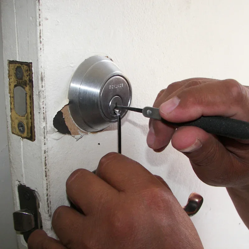 Close-up view of a locksmith working on a residential door lock in Sugar Land, Texas. Two hands are visible working directly on a round silver deadbolt lock mounted on a white-painted wooden door. One hand holds a black-handled locksmith tool inserted into the keyway of the deadbolt, while the other hand steadies a second thin metal tool positioned inside the lock. The silver deadbolt has a circular brushed-metal exterior and a visible key cylinder at its center. The area around the lock shows chipped and damaged white paint, with a section of exposed brown wood beneath the hardware. To the left of the deadbolt is the door strike plate, which has an aged brass-colored finish and visible screw holes, while a separate darker metal latch mechanism is visible lower on the door edge. The close-up composition focuses on the locksmith's hands, tools, deadbolt, door hardware, and the condition of the door around the lock. The image represents residential locksmith service in Sugar Land, Texas, including professional work on residential door locks and locking mechanisms.