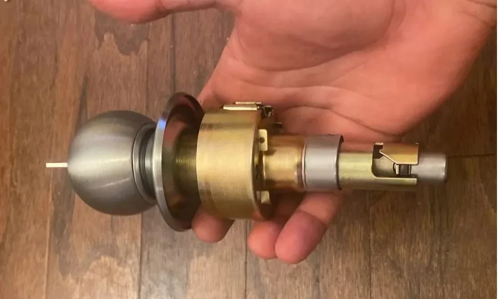 Residential locksmith services in Conroe, TX represented by a disassembled residential door lock and latch assembly being held in a person’s hand above a brown wood-grain floor. The lock hardware is shown horizontally from the side, with a rounded silver metal door knob on the left, a dark metallic circular mounting plate behind the knob, and a large brass-colored cylindrical lock body in the center. The brass lock housing connects to a silver metal cylindrical latch mechanism extending toward the right, ending in a rectangular latch component with a visible internal opening and a small brass-colored section near the top. Several connecting rings, collars, and metal components are visible between the knob and latch body, showing the internal structure of the residential door-lock assembly. The person’s fingers support the lock from underneath while the palm and other fingers are visible behind the hardware. The contrasting silver, brass, and dark metallic finishes of the lock components are clearly visible against the warm brown wooden floor. The image illustrates residential locksmith services in Conroe, TX.