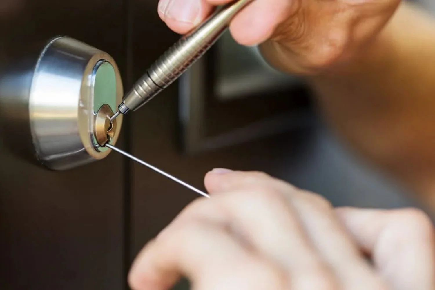 A close-up view of a locksmith working on a residential. The image focuses on a round silver-colored cylindrical door lock mounted on a dark brown door. The lock has a brass-colored key cylinder in the center, with two thin metal locksmith tools inserted into the keyway. One hand is holding a silver-colored, textured metal lock-picking tool positioned inside the upper part of the keyway, while the other hand holds a thin metal tension tool inserted into the lower portion of the lock. The locksmith’s fingers and hands are visible around the tools, carefully working on the internal locking mechanism. The silver lock housing contrasts with the dark brown door, while the brass-colored cylinder is clearly visible at the center. The background is intentionally out of focus, keeping the lock, tools, and locksmith’s hands as the main subjects. The image represents residential locksmith services in Kingwood, Houston, Texas, showing professional work on a residential door lock and locking mechanism.