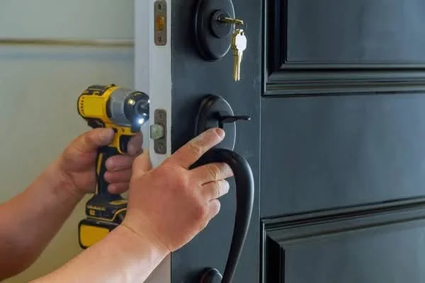 A close-up view of a locksmith working on a dark gray residential front door in League City, Texas. The locksmith is using a yellow-and-black cordless power drill with an attached work light near the door’s lock hardware, while the other hand is positioned on the black curved door handle and lock area. The door is open slightly, revealing its light-colored edge and several metal latch components along the vertical edge. A round black deadbolt lock is installed near the upper portion of the door, with a brass-colored key inserted into the lock and a small key ring hanging below it. Below the deadbolt is a black lock and handle assembly, with the locksmith’s fingers positioned around the locking hardware. The dark gray door has raised rectangular panel detailing visible on the right side. The background is an indoor light-colored wall or doorway area, while the main focus remains on the locksmith’s hands, power drill, door locks, handle, and related hardware. The image shows professional residential locksmith work on a home’s door locking system and represents residential locksmith services in League City, Texas.