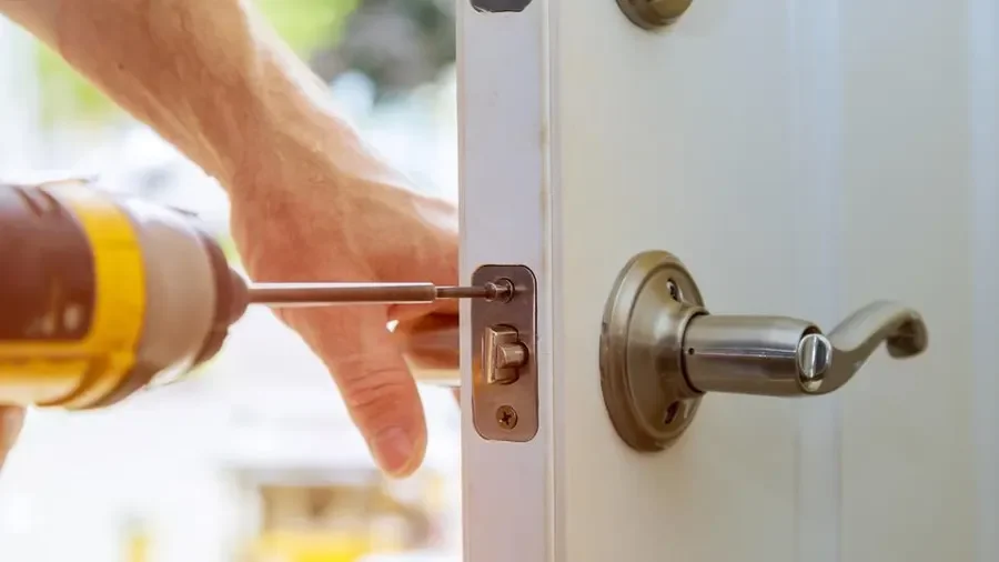 A close-up photograph of a residential locksmith working on the lock hardware of a white exterior-style door in Pearland, TX. The right side of the image shows the white door with a brushed-metal lever handle mounted on a round metallic backplate, along with additional circular lock hardware visible higher on the door. At the edge of the door, a rectangular metal lock plate contains the lock mechanism, with a yellow-and-black cordless power drill inserted directly into the lock cylinder. On the left side, a person’s hand and forearm are visible holding a yellow-and-black cordless power drill fitted with a long metal bit that is positioned toward the lock cylinder. Another hand is visible behind the tool near the door, appearing to holding the door handle. The lock hardware has metallic bronze, brass, and silver tones, contrasting against the plain white door surface. The background is softly blurred with bright natural daylight, green foliage, and light-colored outdoor surroundings visible through the shallow depth of field. The photograph is tightly framed around the locksmith’s hands, power tool, lock cylinder, and door hardware, emphasizing hands-on residential lock service work.