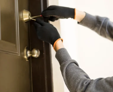 A close-up view of a residential locksmith working on a dark brown wooden front door in The Woodlands, Texas, after the homeowner’s key was lost. The locksmith is using specialized lock-picking tools to open the door’s upper deadbolt lock, with one tool inserted into the brass-colored key cylinder and another tool positioned alongside it. The locksmith is wearing black protective gloves with orange trim and a gray long-sleeve shirt. Only the locksmith’s arms and hands are visible in the image. The door has a brass-colored deadbolt near the upper portion and a matching round brass-colored door knob below it. The dark brown door features raised rectangular panel detailing, while the surrounding area is a light-colored interior or doorway space. The image focuses closely on the locksmith’s hands, lock-picking tools, deadbolt, and residential door hardware, showing professional lockout assistance for a homeowner who no longer has their original key. The image represents residential locksmith services in The Woodlands, Texas.